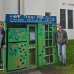 Photograph of two people standing on either side of an outdood refrigerator in front a a church labeled "St. Joseph Parish"