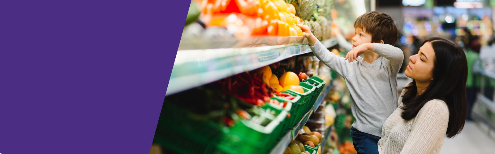 Stock photo of a mom holding child to reach for vegetables on a high shelf at the grocery store